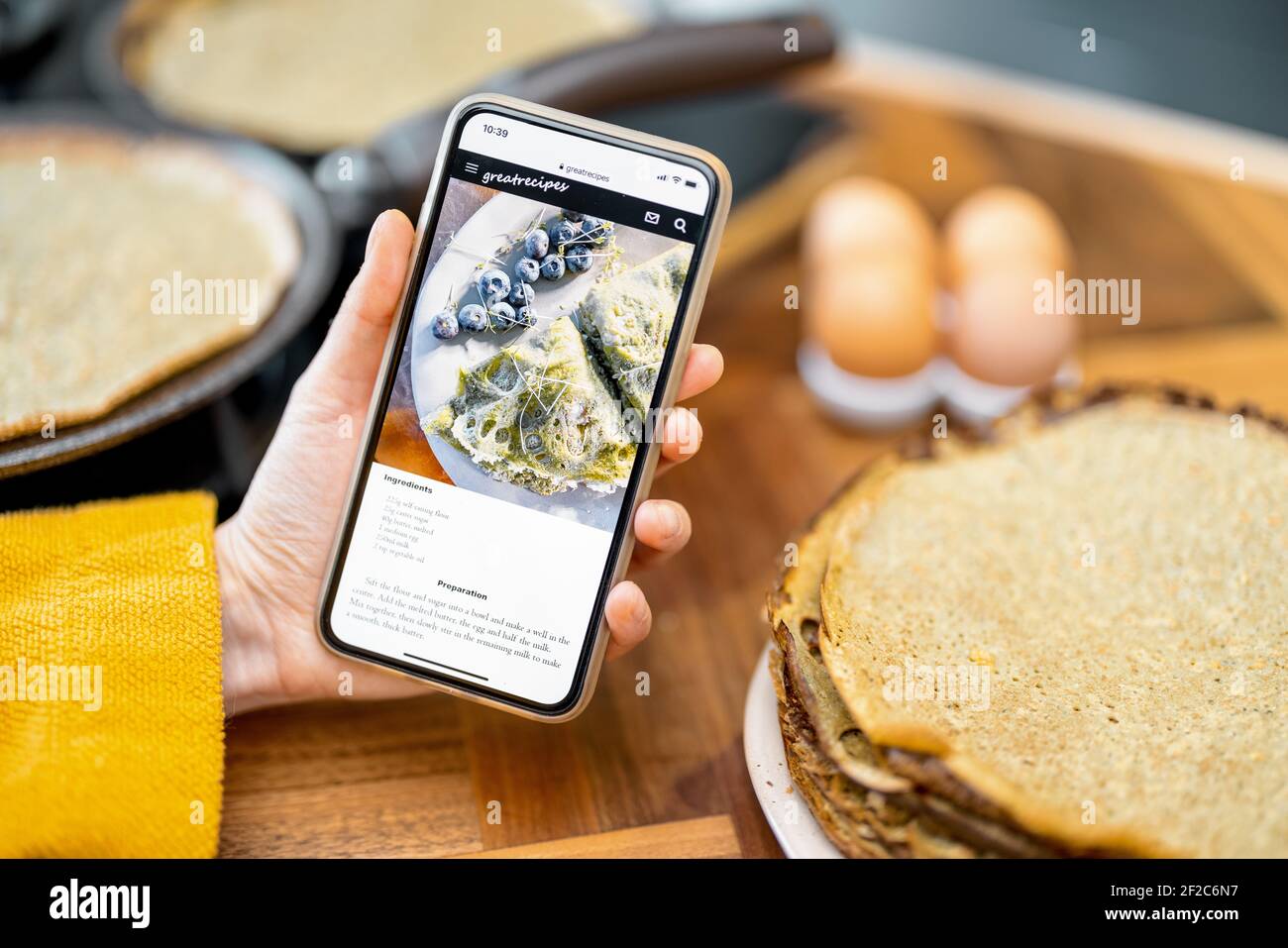 Tenere smartphone con la ricetta di frittelle yummy per un colazione con ingredienti alimentari sul tavolo da cucina dietro Foto Stock