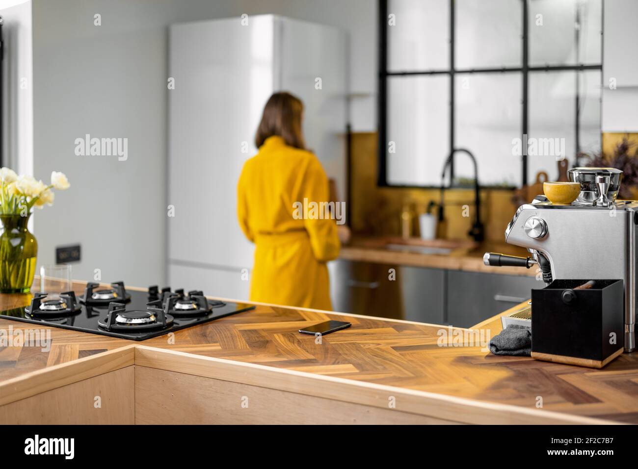 Elegante cucina interna con tavolo in legno e macchina per il caffè. Donna in accappatoio prende il cibo dal frigorifero sullo sfondo. Fuoco anteriore sul piano di tavola di legno e stufa nera. Foto Stock