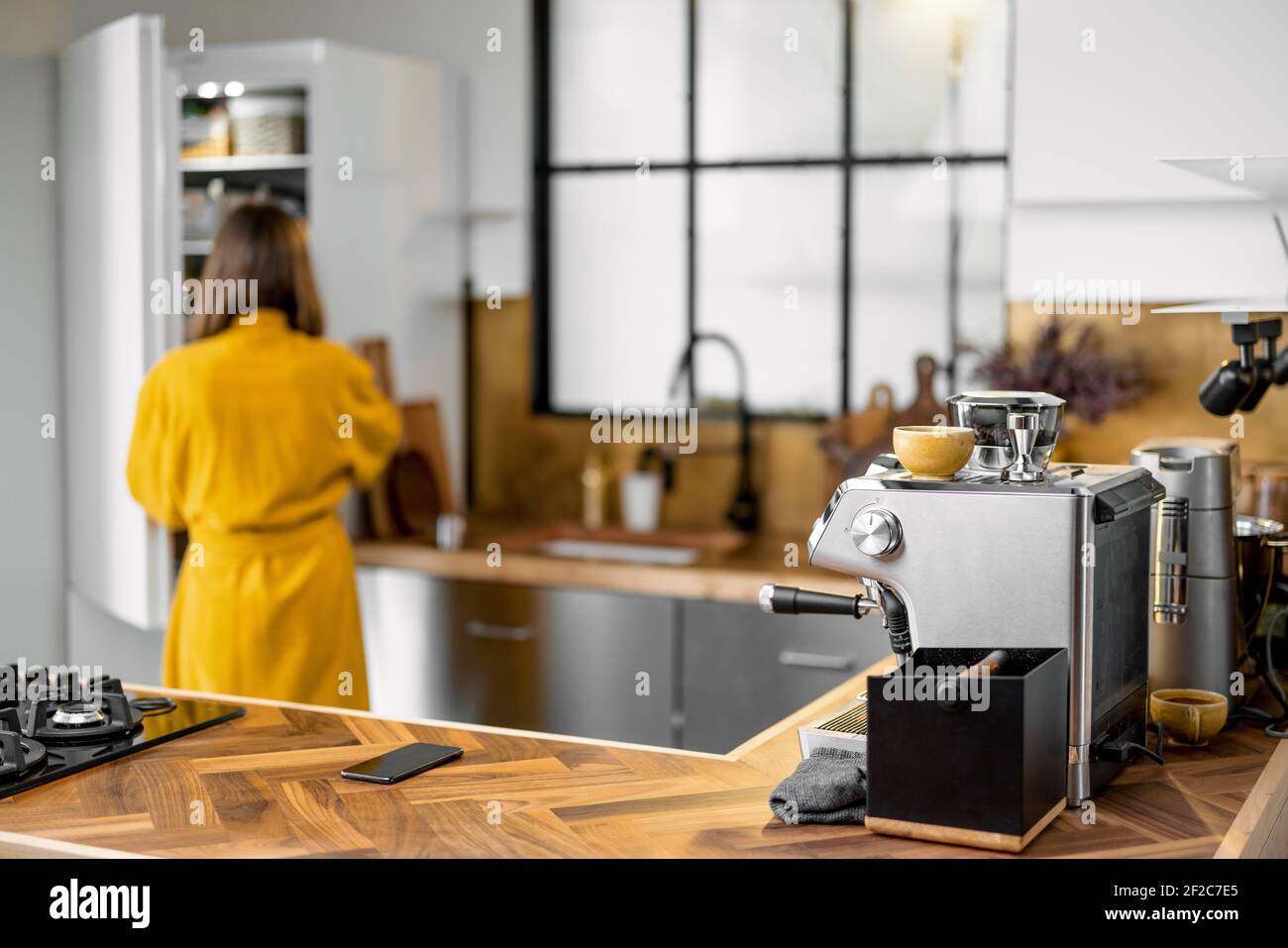Elegante cucina interna con tavolo in legno e macchina per il caffè. Donna in accappatoio prende il cibo dal frigorifero sullo sfondo. Messa a fuoco frontale sul piano di legno. Foto Stock
