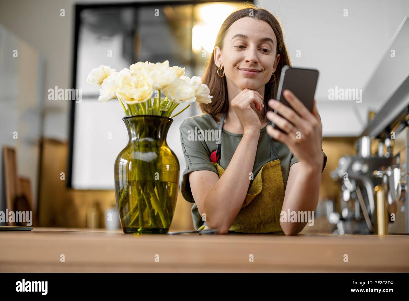 Giovane casalinga in grembiule verde ordinando con smartphone un bouquet di tulipani freschi. Fiorista con fiori in cucina a casa. Foto Stock