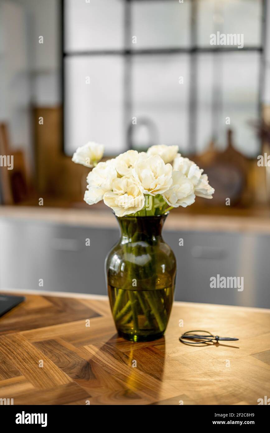 Vaso con fiori bianchi su elegante fondo interno cucina con tavolo in legno. Concetto di intimità domestica. Bouquet di tulipani in vaso di vetro verde. Foto Stock
