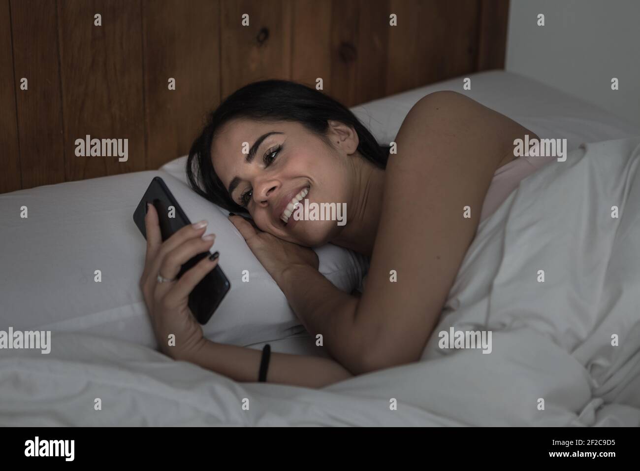 Contenuto donna che si trova in letto sotto coperta di sera e. lettura dei messaggi sul telefono cellulare Foto Stock