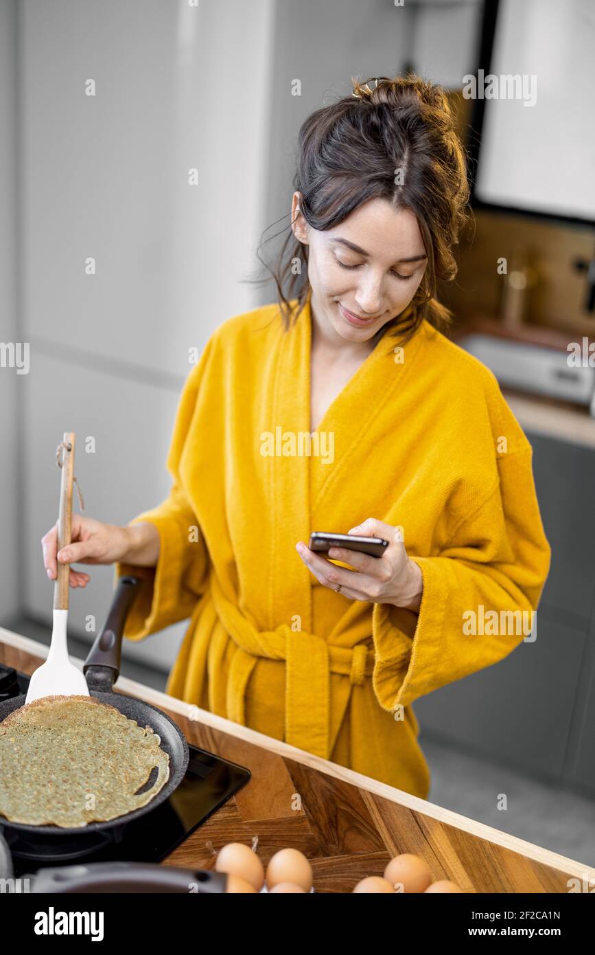 Donna felice vestita in accappatoio cuoce frittelle per la colazione, guarda sul telefono mentre si trova vicino al piano cottura nella cucina moderna a casa Foto Stock