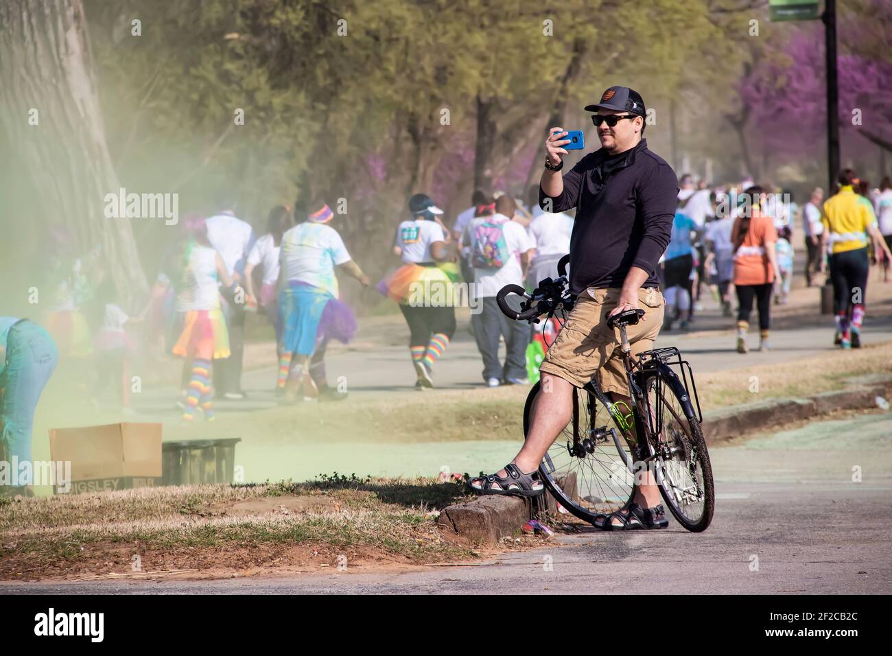 4-6-2018 Tulsa USA - uomo in bicicletta con cappello da baseball e i pantaloncini da carico e gli occhiali da sole si fermerono per scattare foto al telefono di partecipanti in holi colore correre con Foto Stock