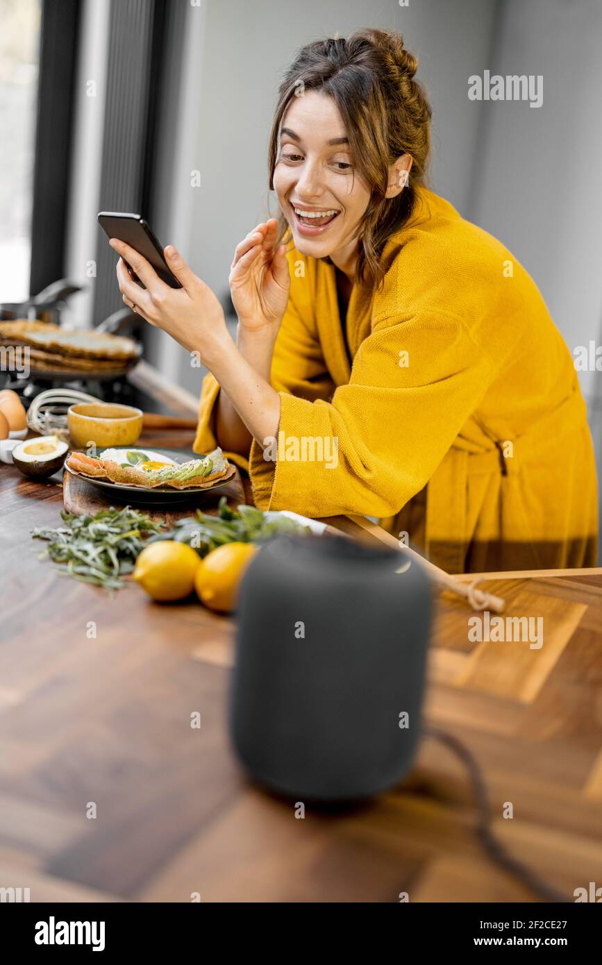 Donna felice in accappatoio che controlla i dispositivi domestici con comandi vocali, parlando a una colonna intelligente durante la colazione in cucina a casa. Concetto di casa intelligente Foto Stock
