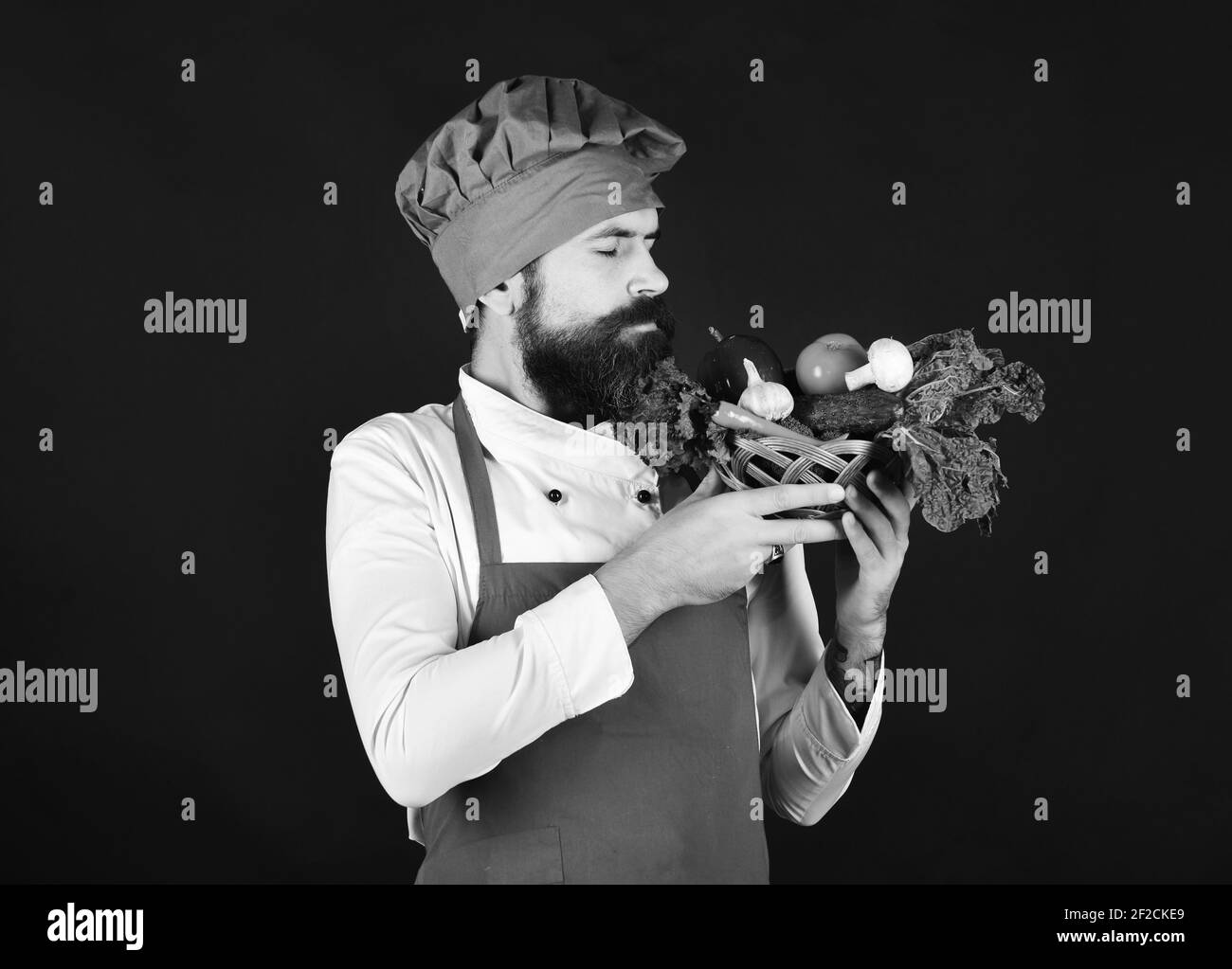 Lo chef conserva lattuga, pomodoro, pepe e funghi. Cuocere con il viso sognante in borgogna uniforme odori verdure in vaso di vimini. Concetto di drogheria verde. Uomo con barba su sfondo nero. Foto Stock