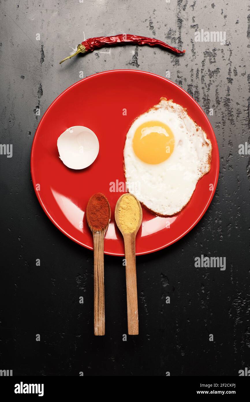 La frittata, guscio d'uovo e riflesso rendere volto sorridente. Piatto con uovo fritto vicino a cucchiai con spezie e peperoncino rosso. La colazione e il cibo arte concetto. Alimenti posti sulla piastra di colore rosso su sfondo nero, vista dall'alto Foto Stock