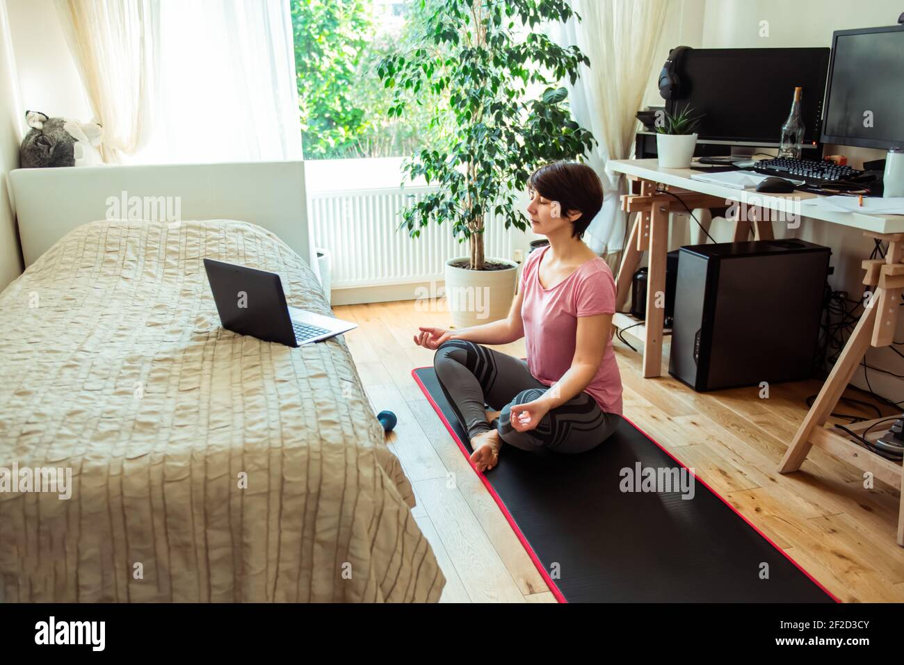 Una donna seduta su un tappetino di fronte al letto mentre fa yoga posizione in camera da letto. Relax yoga pratica a casa. Stile di vita sano. Workou in linea domestico Foto Stock