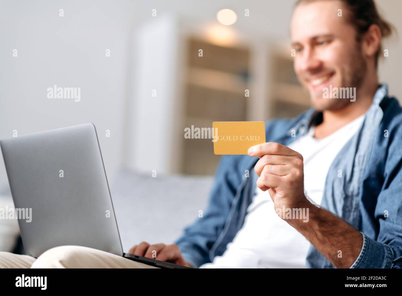 Ragazzo caucasico sorridente defocused, in abiti casual di stile, si siede sul divano a casa, tiene un laptop e una carta bancaria, effettua il pagamento online per acquisti o bollette, inserisce un numero di carta Foto Stock