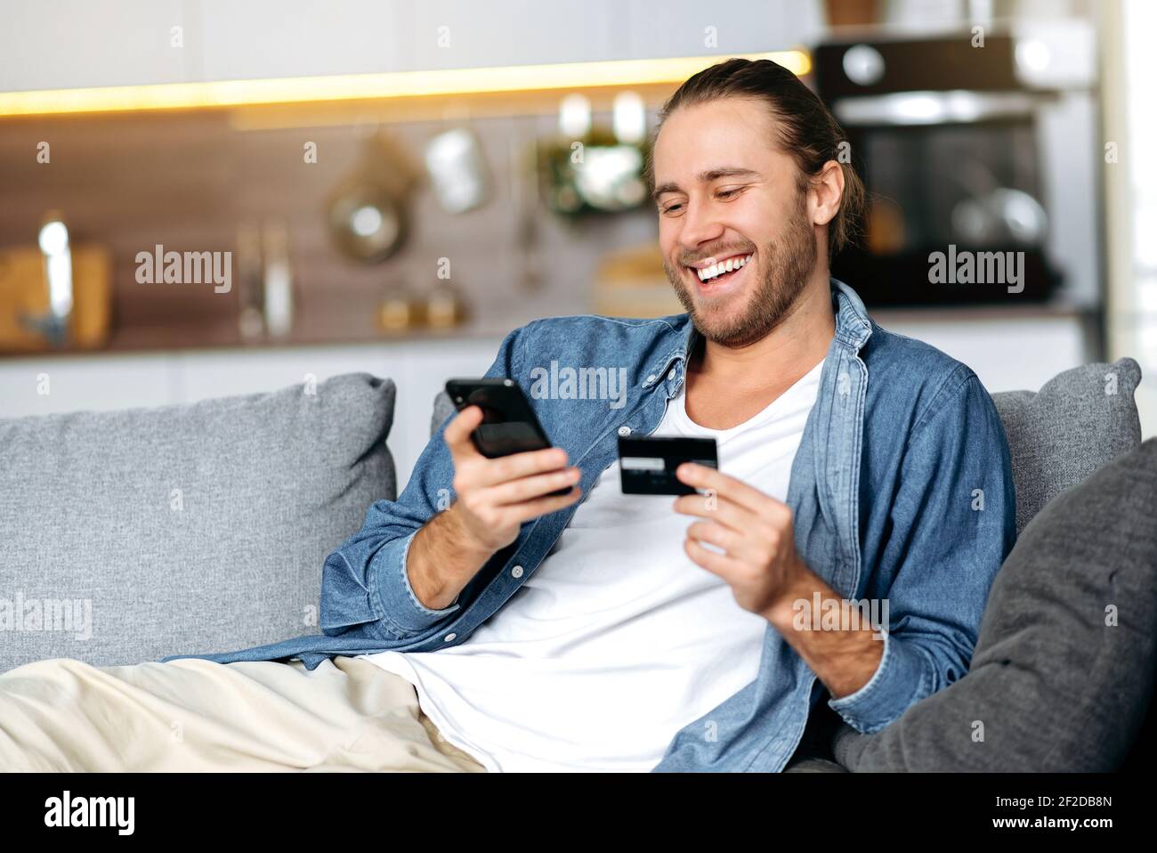 Un ragazzo caucasico sorridente felice, in abiti casual di stile, si siede sul divano a casa, tiene un telefono e una carta bancaria in mano, effettua il pagamento online per gli acquisti, inserisce un numero di carta Foto Stock