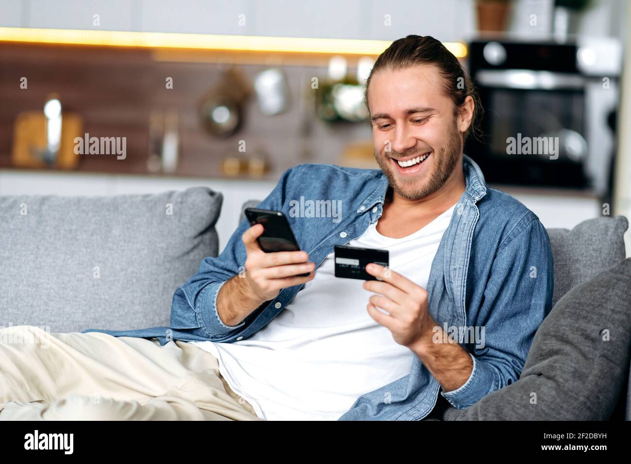Un ragazzo caucasico sorridente soddisfatto, in abiti casual di stile, si siede sul divano a casa, tiene un telefono e una carta di credito nelle sue mani, paga per gli acquisti online, inserisce un numero di carta, shopping online Foto Stock