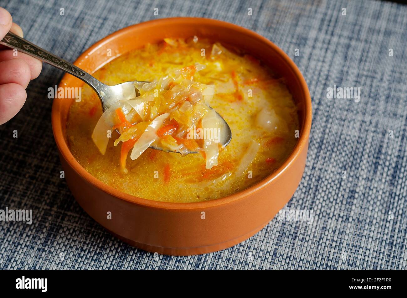 Una mano con un cucchiaio di metallo mescola la panna acida in un recipiente di terracotta marrone con zuppa di cavolo. Una porzione di zuppa pronta su una tovaglia grigio-blu. Cl Foto Stock
