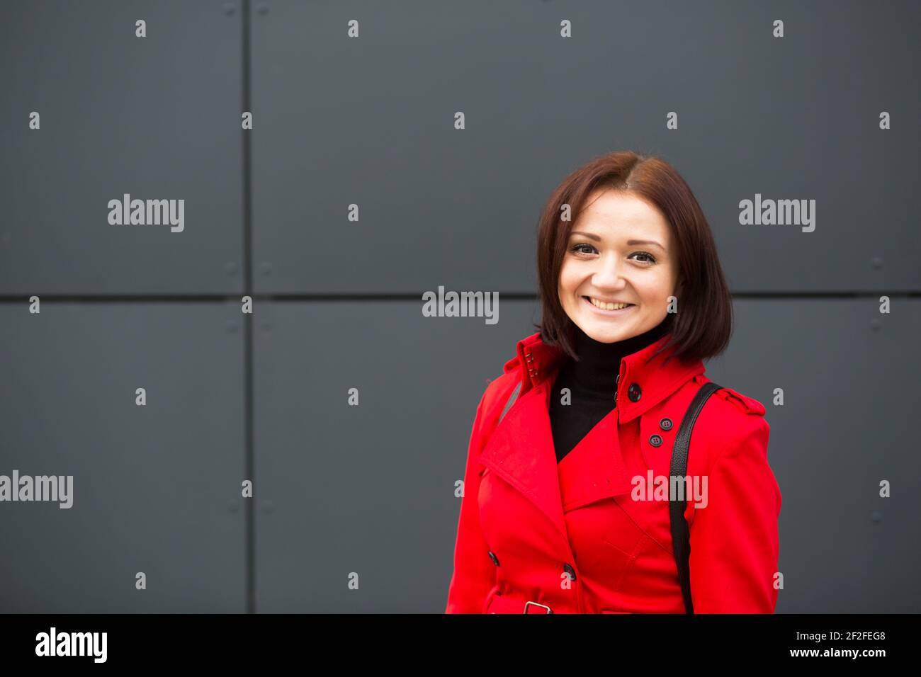 Una giovane e felice donna caucasica in un impermeabile rosso con uno zaino in pelle nera contro una parete grigia. Stile di vita, emozioni, femminilità, ritratto Foto Stock