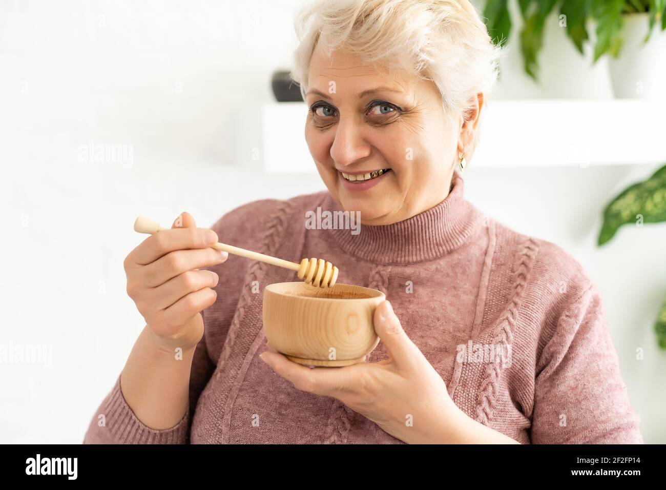 Primo piano ritratto di affascinante donna matura, tenendo il vaso di miele con cucchiaio, guardando la macchina fotografica, isolato su sfondo bianco. Foto Stock