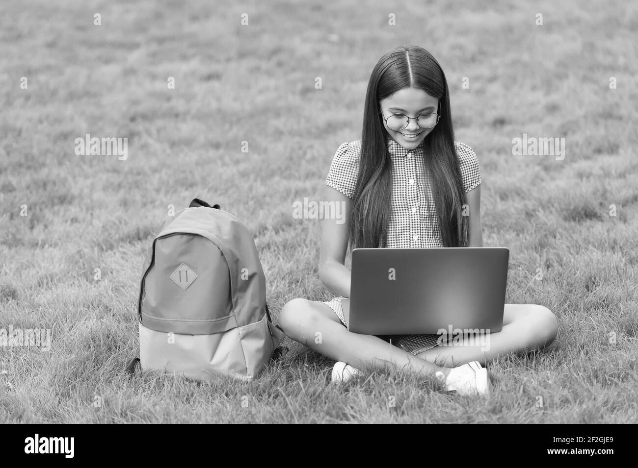 smart happy kid indossare occhiali utilizzando il computer portatile per fare compiti a scuola online seduto in parco su erba verde con zaino, istruzione. Foto Stock