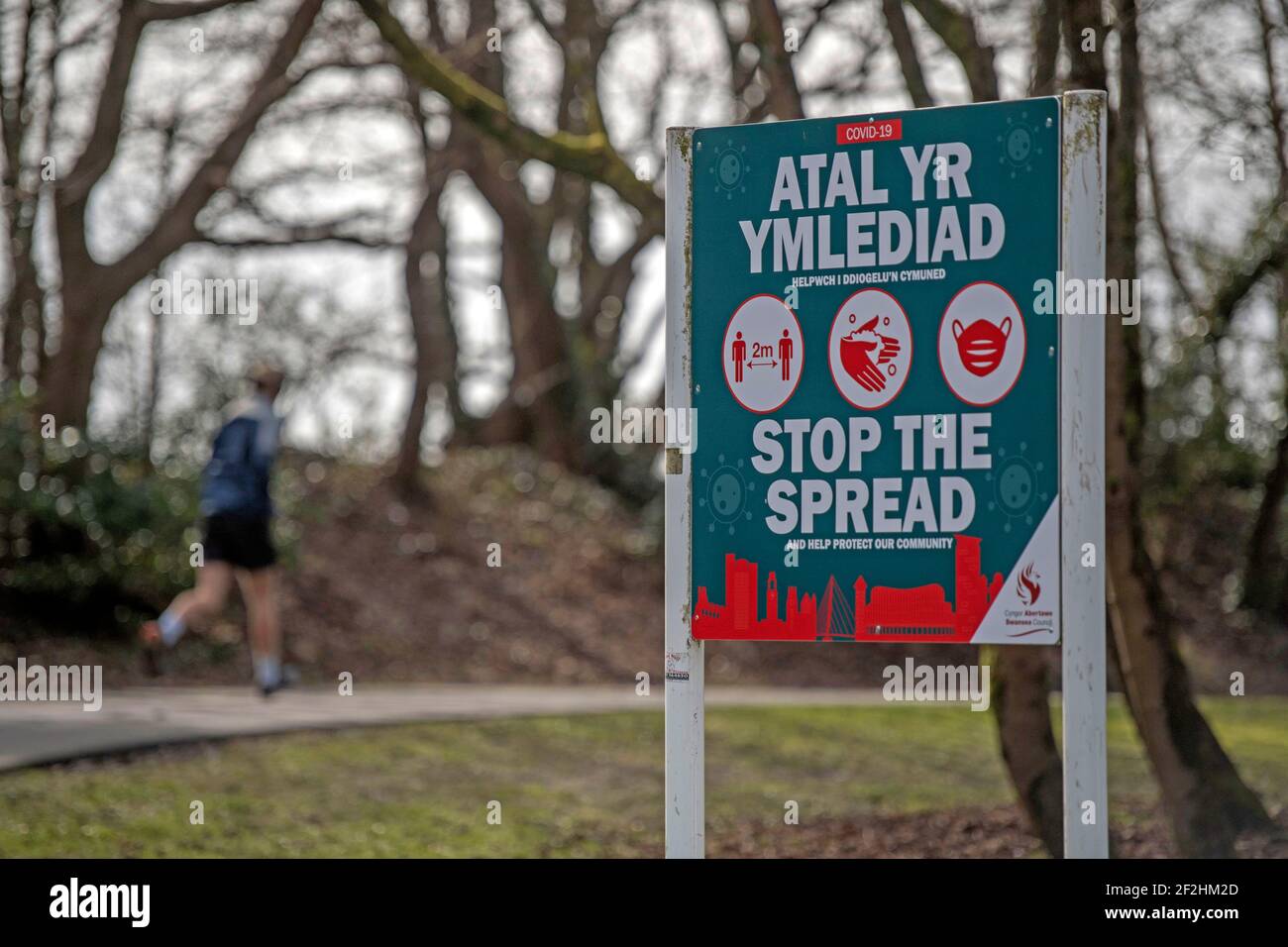 Swansea, Regno Unito. 12 marzo 2021. Un corridore si fa strada oltre un cartello di avvertimento Covid 19 sul sentiero lungomare che corre tra Swansea e il piccolo villaggio Mumbles durante il blocco questo pomeriggio. Il governo gallese ha annunciato oggi che sta iniziando a rilassare le sue regole di blocco. Credit: Phil Rees/Alamy Live News Foto Stock