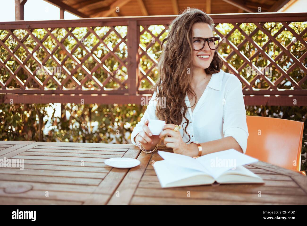 donna elegante felice in camicia bianca con libro e occhiali seduti al tavolo bere caffè nella terrazza dell'hotel della pensione. Foto Stock