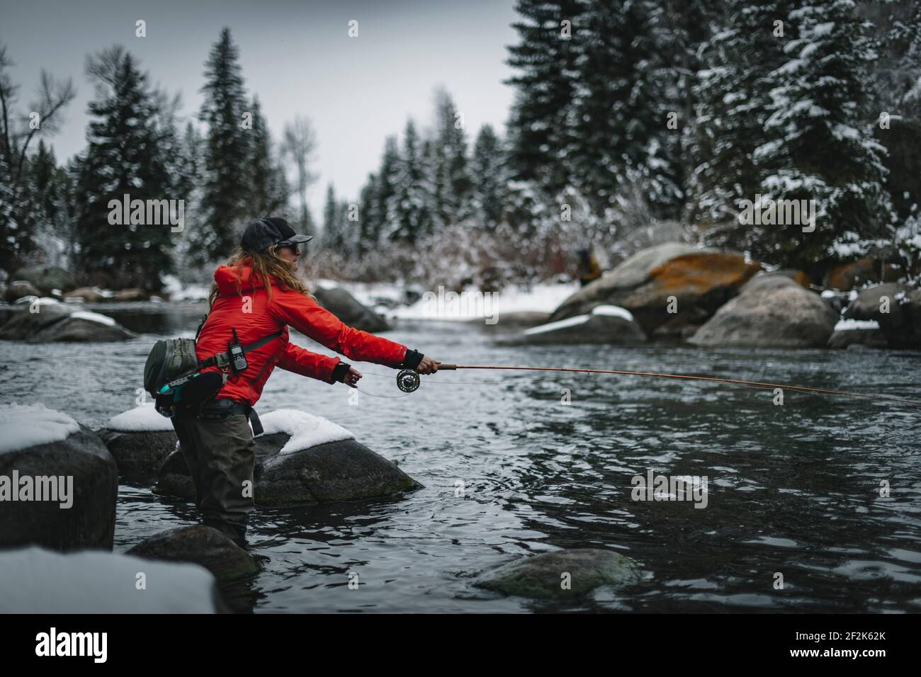 Vista laterale della pesca con mosca femminile in piedi sulla neve roccia coperta dal fiume durante l'inverno Foto Stock