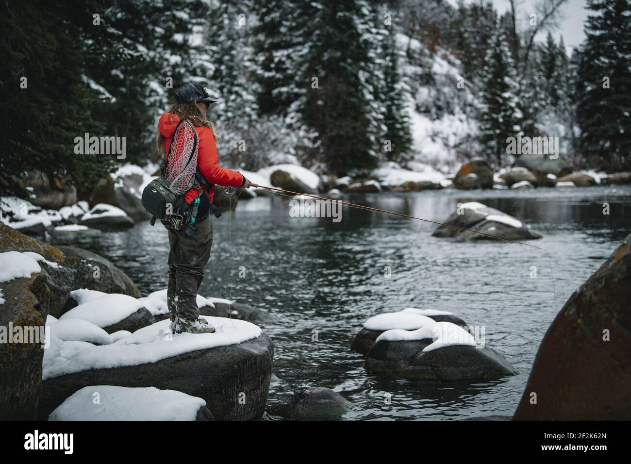 Vista laterale della donna pesca a mosca in piedi sulla neve roccia coperta dal fiume durante l'inverno Foto Stock