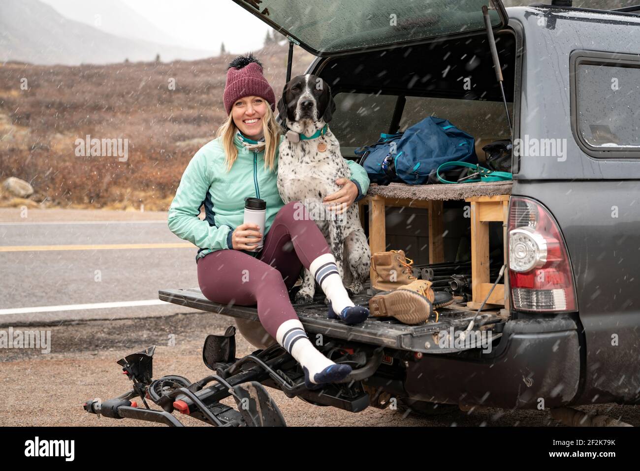 Giovane donna sorridente seduta con il cane in tronco di fuori strada veicolo in caso di nevicata Foto Stock