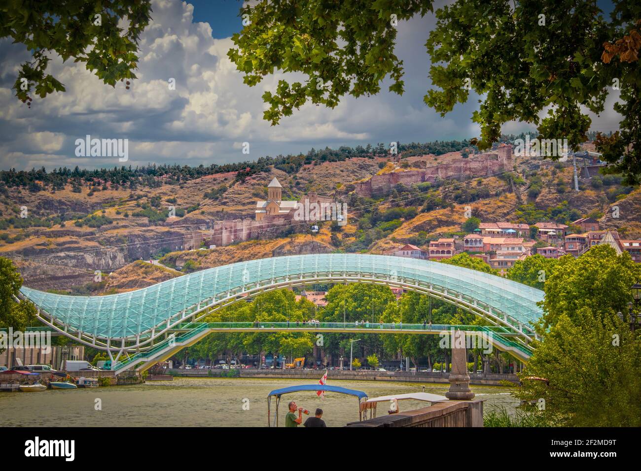2019 07 19 Tbilisi Georgia - Ponte della Pace con l'antica fortezza di Narikala Si affaccia su Tbilisi e si affaccia sulle rive del fiume Mtkvari - Kura con barca Foto Stock