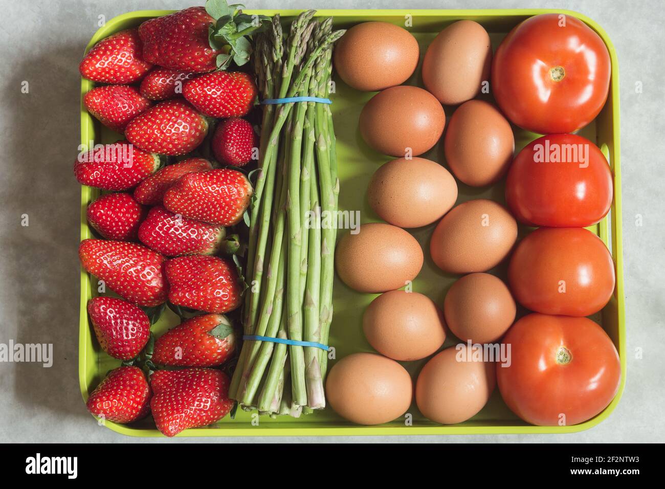 Un vassoio pieno di alimenti biologici e naturali con verdure e frutta. Fragole, asparagi selvatici, pomodori e uova su sfondo verde. Foto Stock