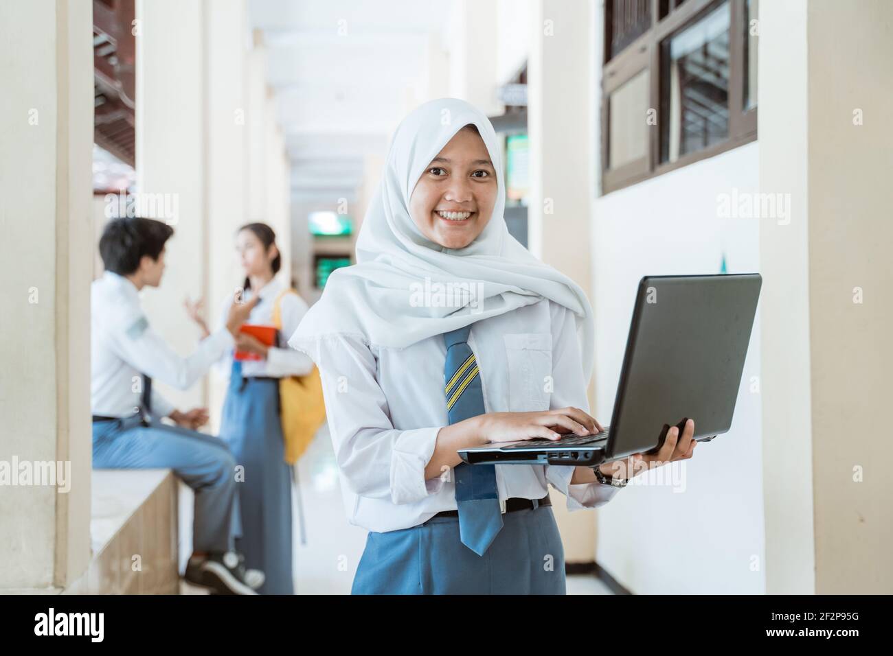 una ragazza in un velo di scuola superiore sorride con un computer portatile sullo sfondo di un corridoio dell'edificio Foto Stock