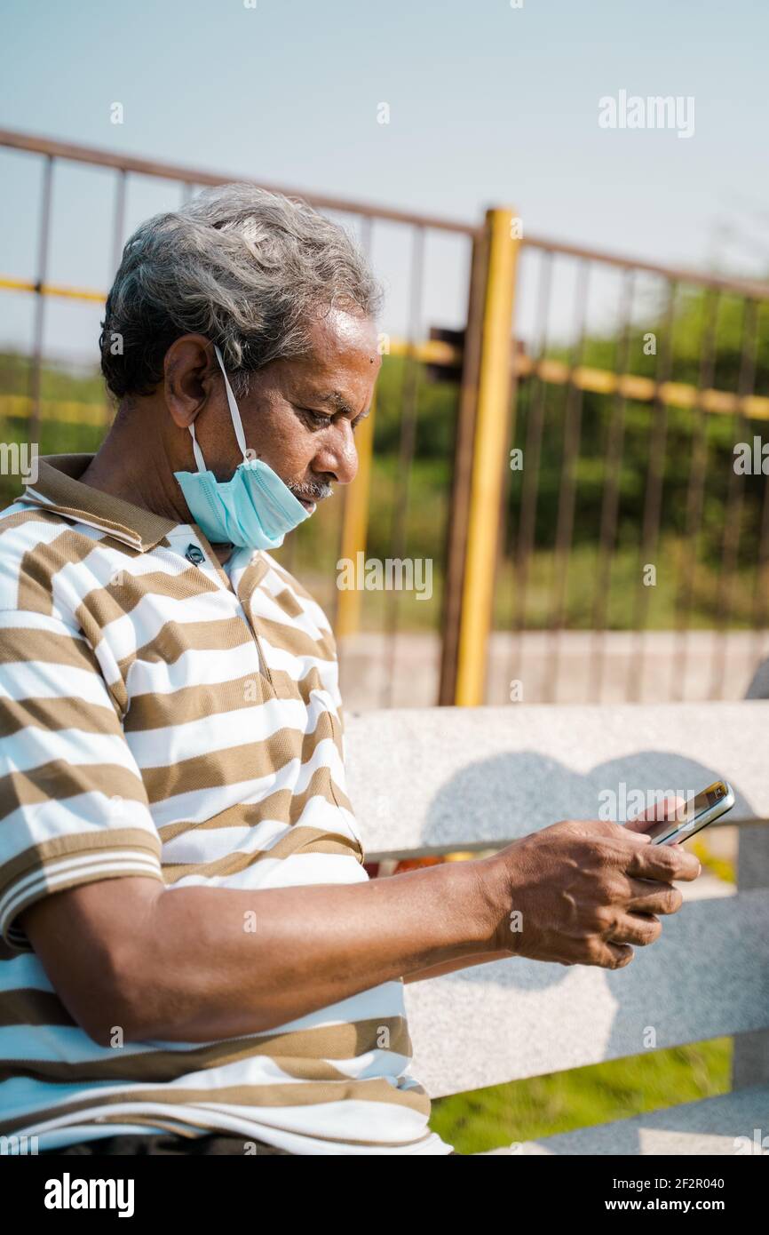 Uomo anziano con maschera medica sotto la mascella utilizzando smartphone al parco - concetto di uso improprio maschera a causa di pandemie coronavirus covid-19. Foto Stock
