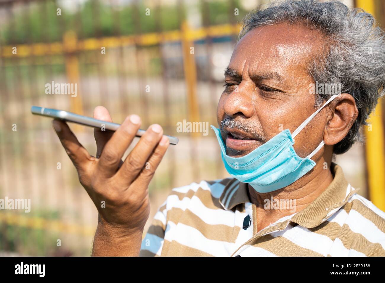 Uomo anziano con maschera medica sotto la mascella che parla sul telefono cellulare al parco - concetto di uso improprio maschera a causa di pandemie coronavirus covid-19. Foto Stock