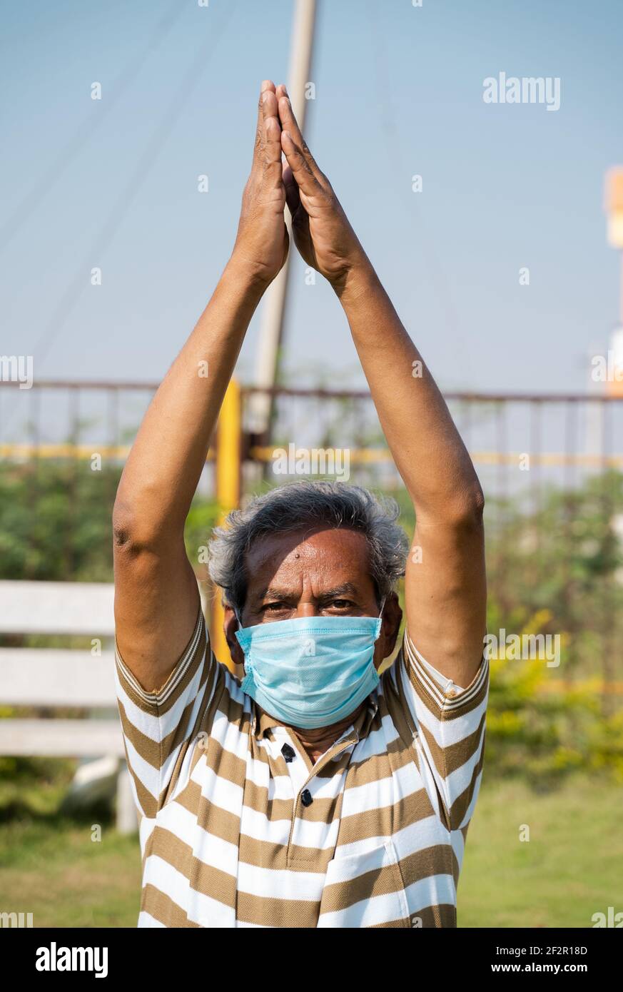 Vecchio uomo con maschera medica occupato facendo surya namaskara yoga o esercizio durante la mattina - concetto di anziani allenamento fitness e assistenza sanitaria Foto Stock