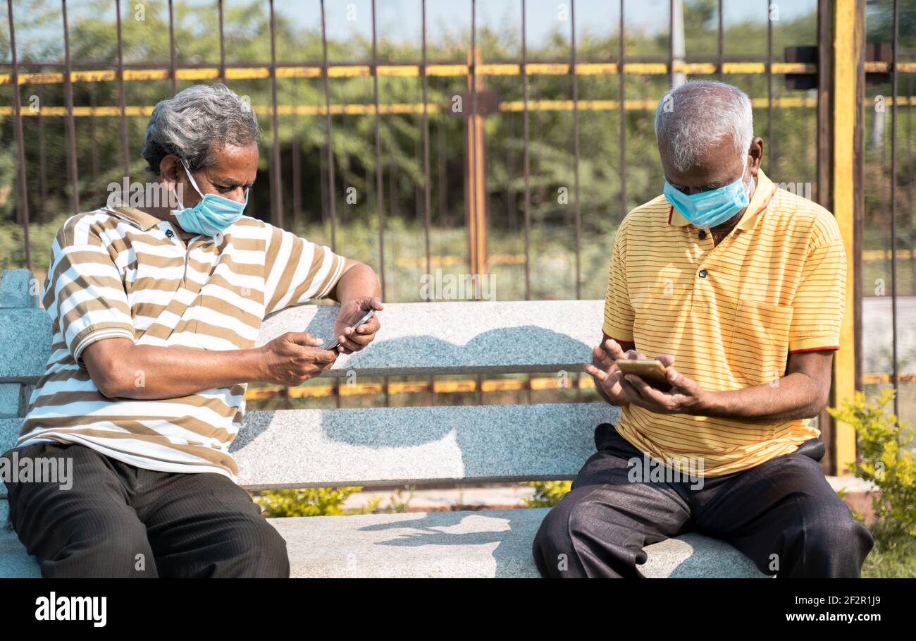 Due persone anziane con maschera medica occupato utilizzando il cellulare telefono mentre si è seduti al parcheggio mentre si mantiene la distanza sociale - concetto anziani uso Foto Stock