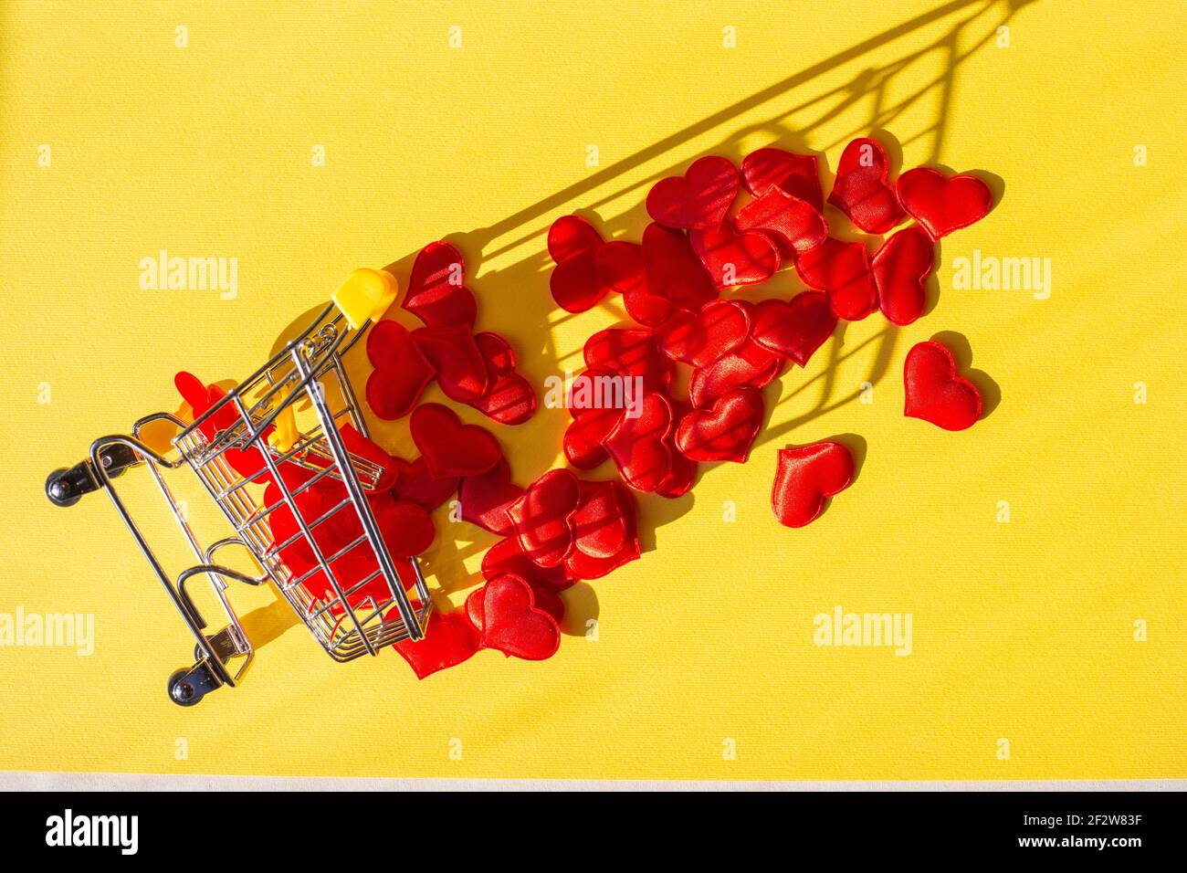 il carrello dello shopping si capovolse e i cuori rossi caddero fuori da esso, uno sfondo giallo. Acquista concetto di amore, flatlay. Foto Stock