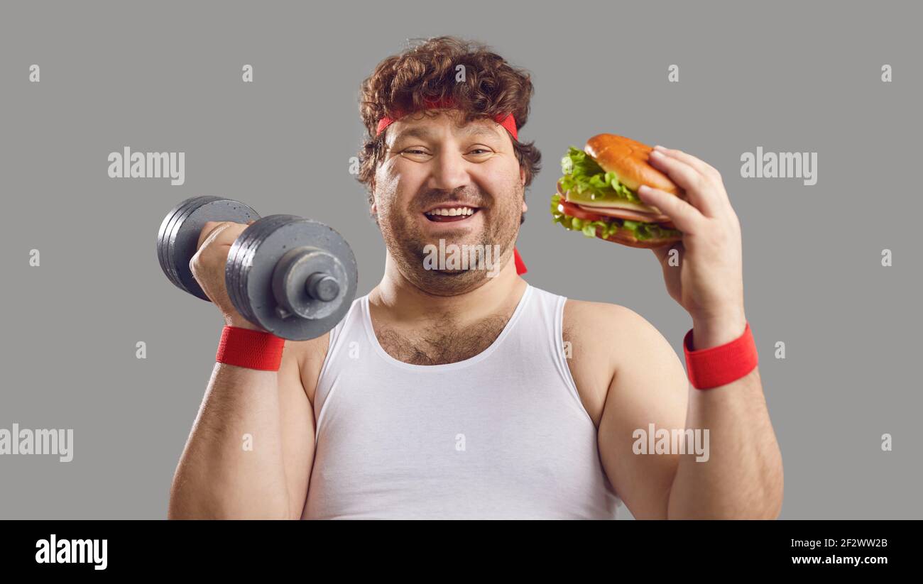 Divertente uomo chubby che tiene dumbbell, mangiando grande hamburger yummy e sorridendo alla macchina fotografica Foto Stock