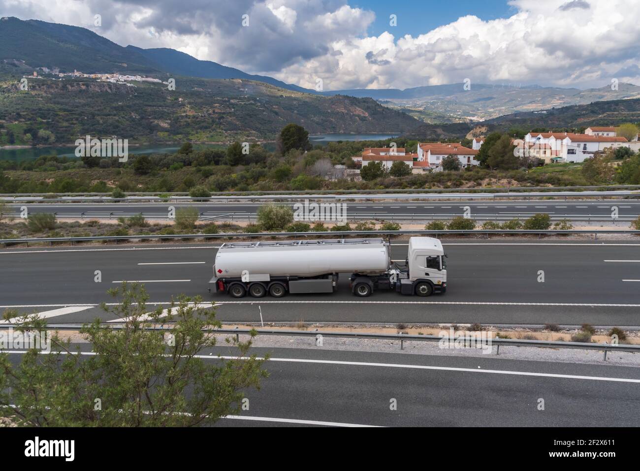 Camion cisterna che guida sulla strada con un paesaggio di montagne, nuvole, un serbatoio e una città sullo sfondo. Foto Stock