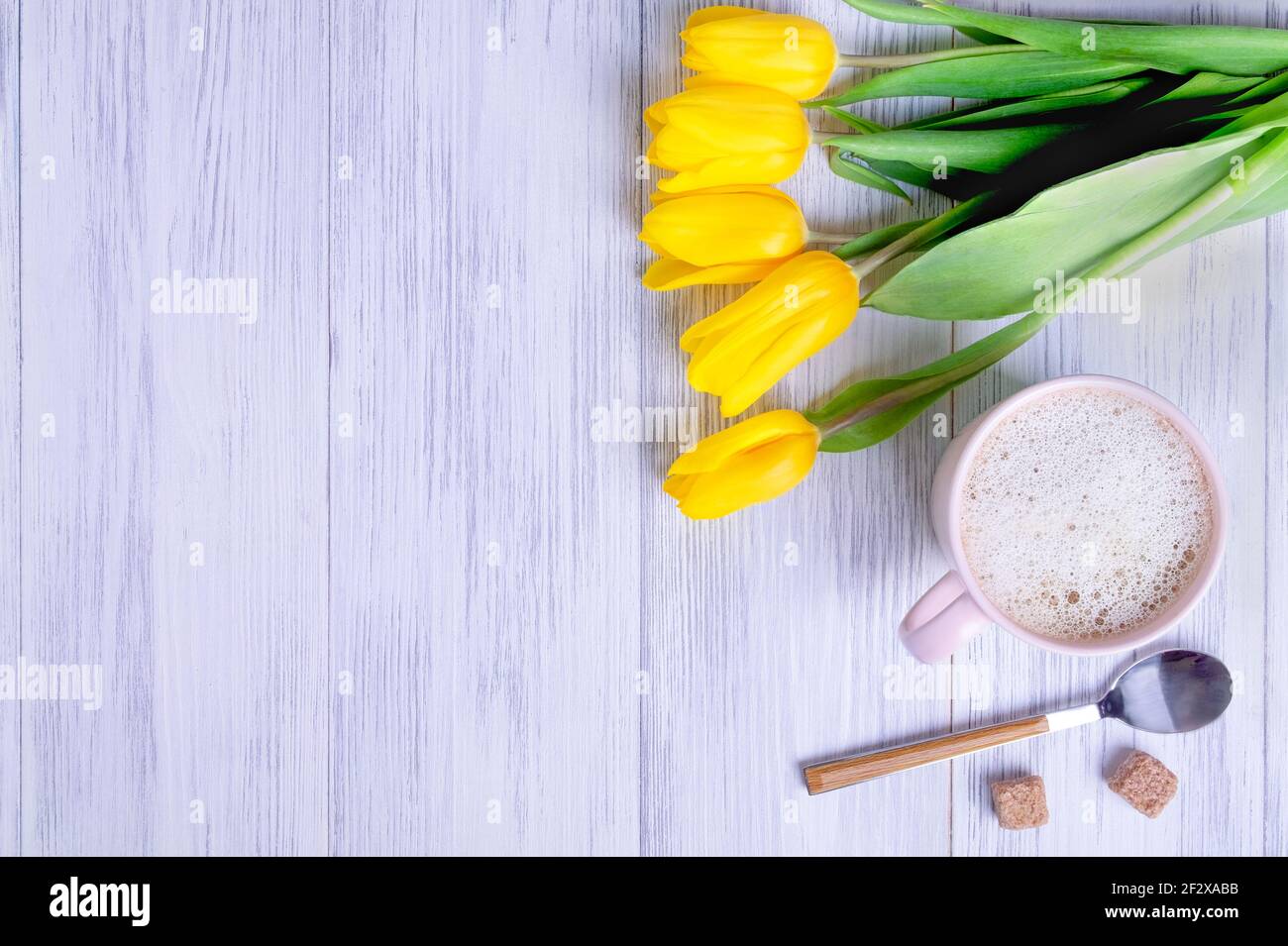 Vista dall'alto di una composizione di un bouquet di tulipani gialli, una tazza rosa con cappuccino con un cucchiaio e zucchero su uno sfondo di legno chiaro. Messa a fuoco selettiva Foto Stock