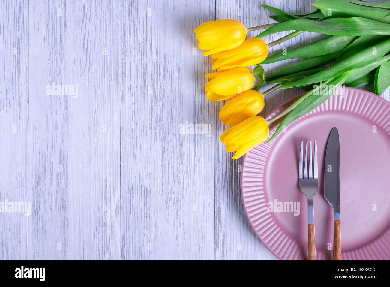 Vista dall'alto di una composizione di un bouquet di tulipani gialli, una piastra rosa con posate su uno sfondo di legno chiaro. Messa a fuoco selettiva. Spazio di copia per il te Foto Stock