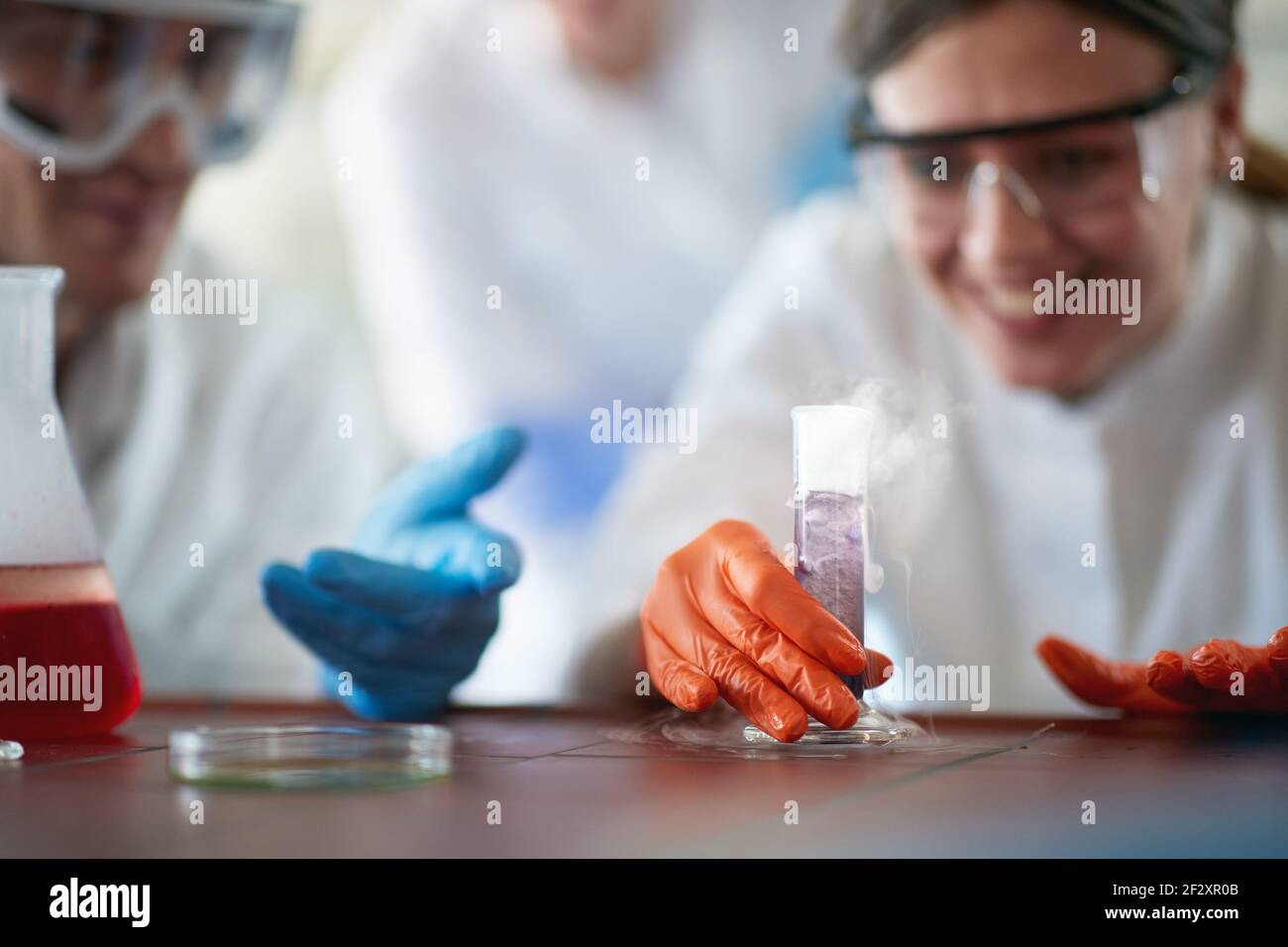 I giovani studenti di chimica che si trovano in un equipaggiamento protettivo godono di una pericolosa reazione chimica in un'atmosfera di lavoro nel laboratorio universitario. Scienza Foto Stock