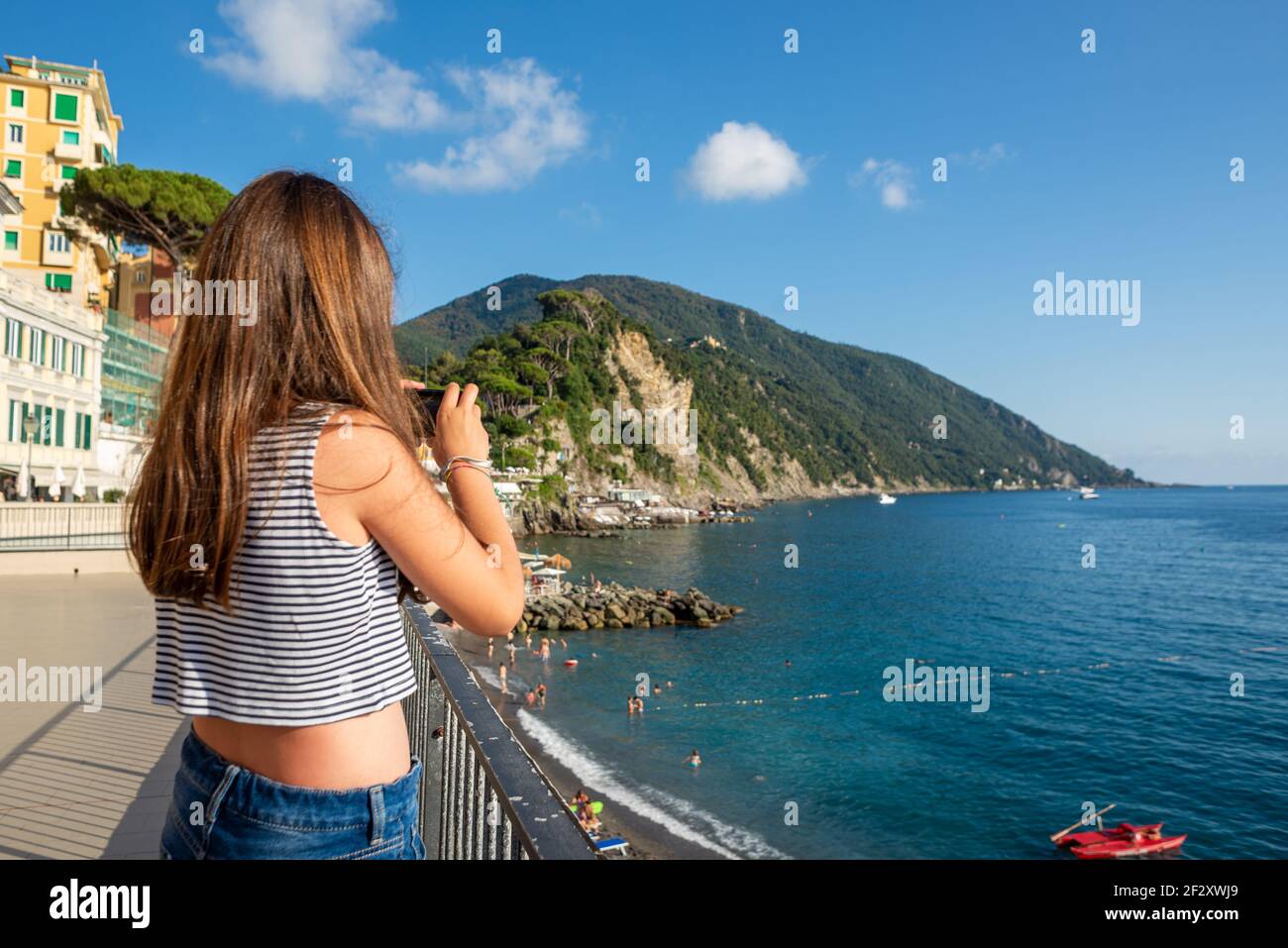 La giovane ragazza dai lunghi capelli marroni sta scattando una foto del mare ligure da una terrazza nella città di Camogli. Ragazza vista da dietro indossando un canotta top Foto Stock