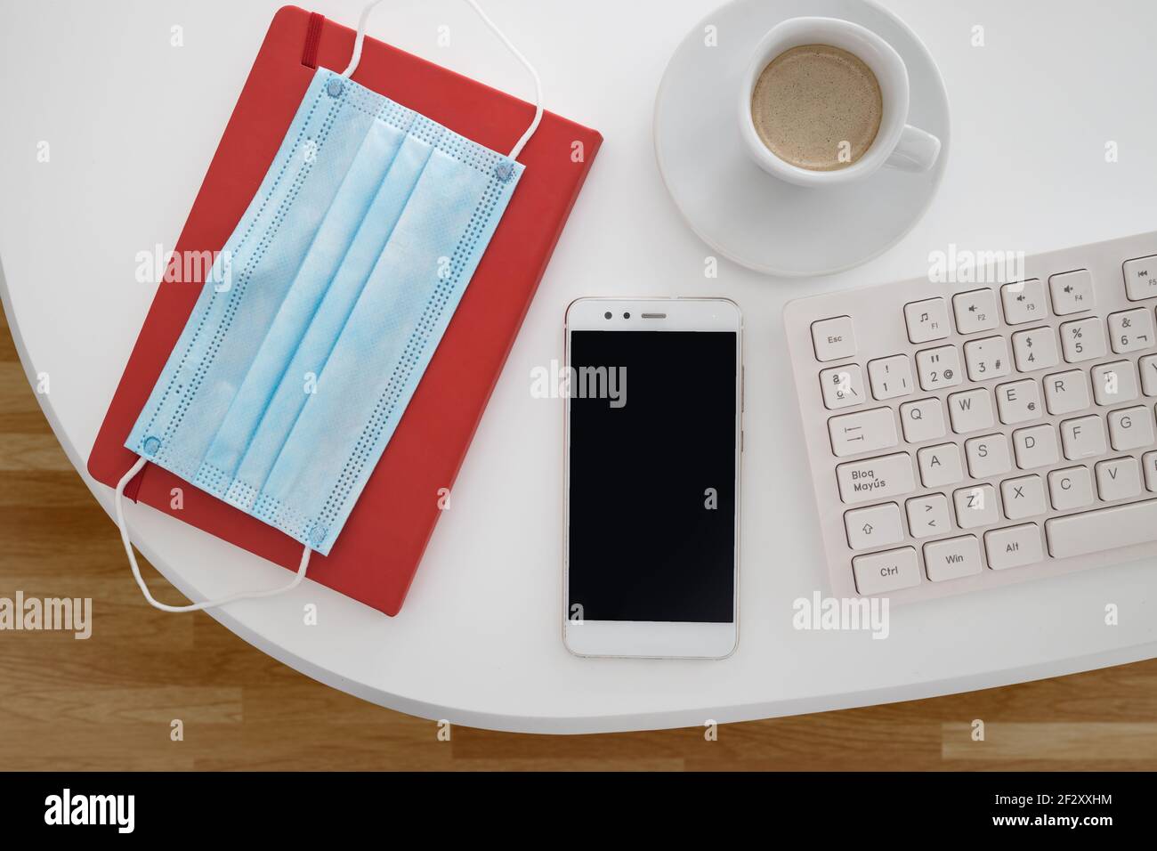 Vista dall'alto della maschera medica posizionata sul notebook e disposta con smartphone e tastiera vicino a una tazza di caffè su bianco scrivania Foto Stock