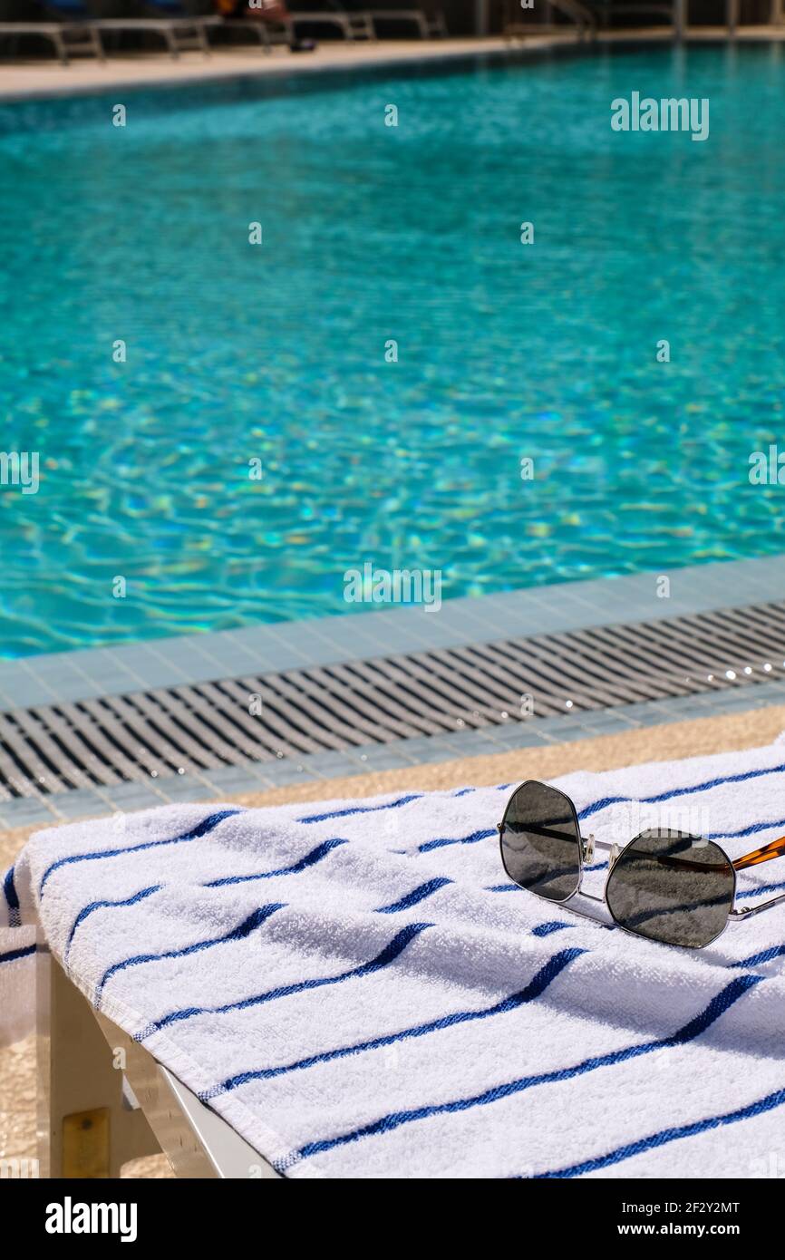 Un paio di occhiali da sole su un asciugamano da piscina bianco con Blue Strips accanto a una piscina in un giorno di sole Foto Stock