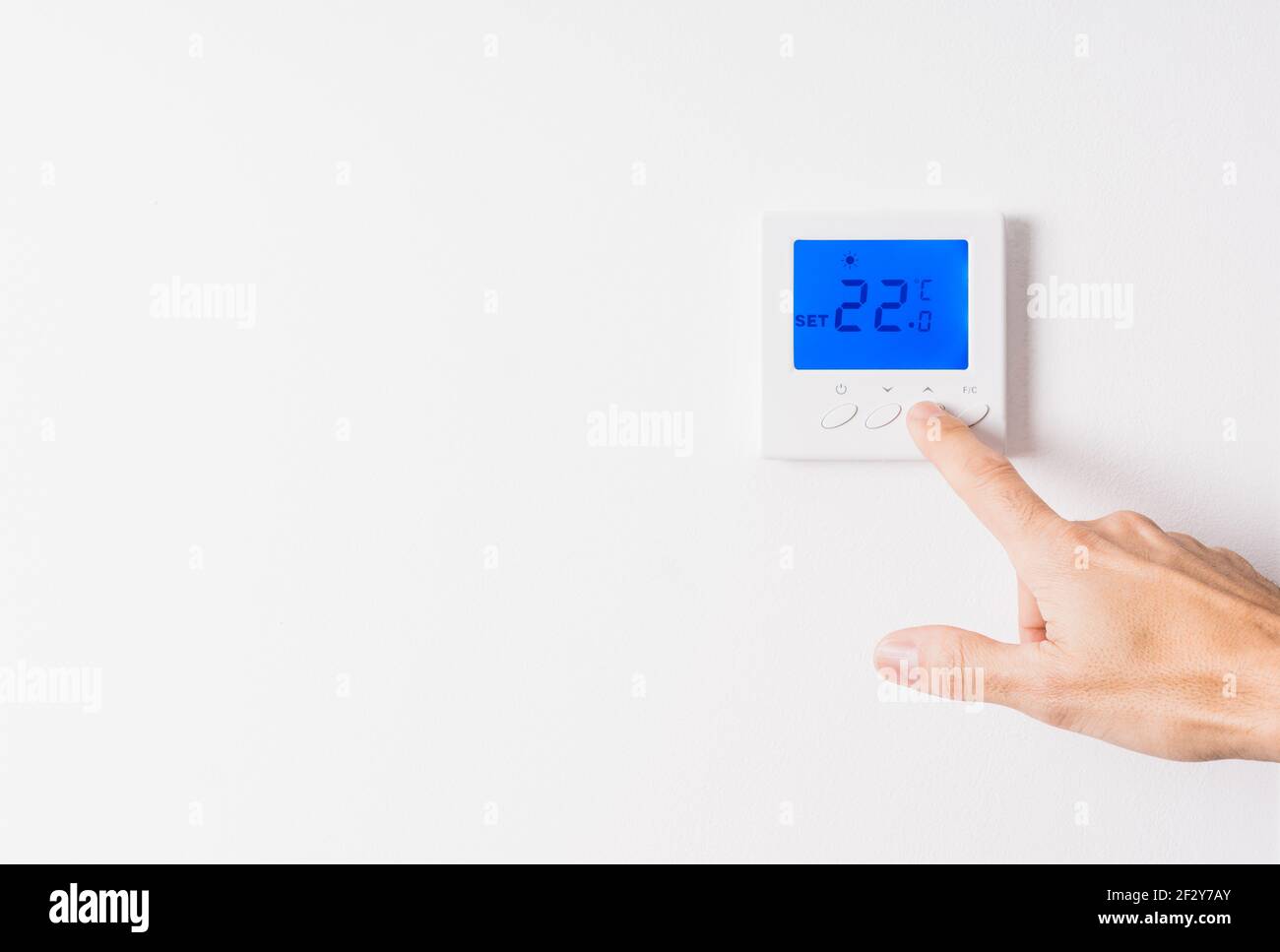 Mano dell'uomo caucasico che regola la temperatura di un termostato bianco a ventidue gradi con il display retroilluminato blu su un parete bianca Foto Stock