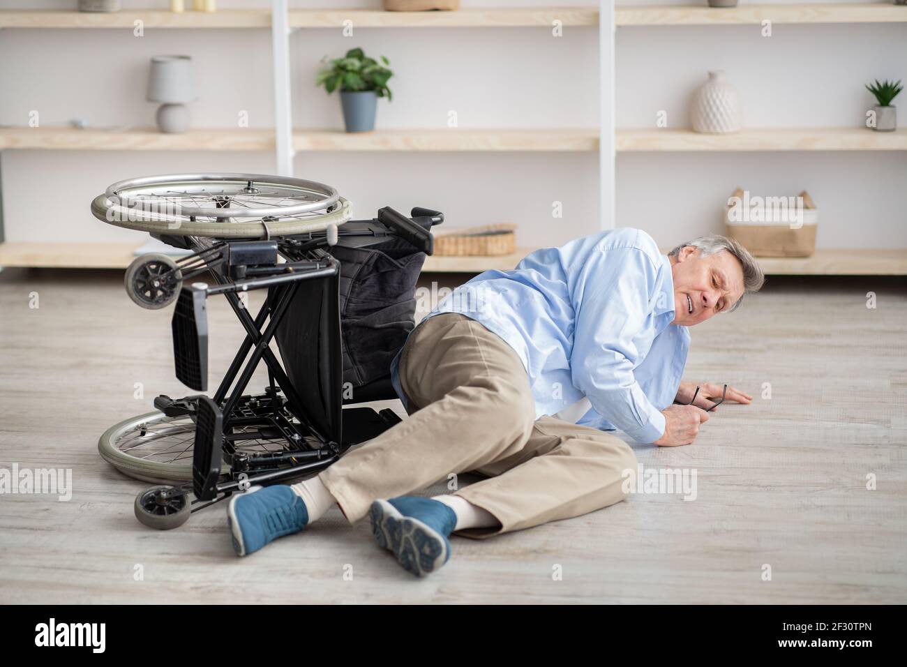 Uomo anziano con disabilità fisica sdraiato sul pavimento dopo la caduta giù dalla sua sedia a rotelle a casa Foto Stock