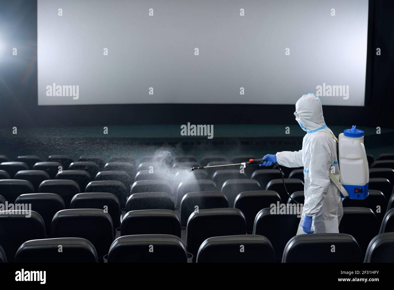 Uomo o donna in indumenti anti-virus protettivi sedili di pulizia in sala cinema con disinfettanti. Concetto di preparazione sala cinema per nuovi visitatori. Foto Stock