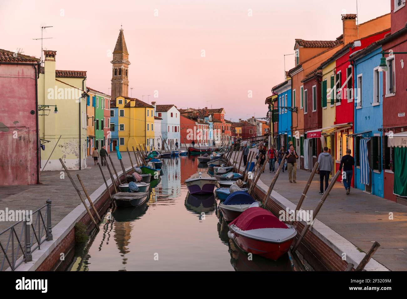 L'isola veneziana di Burano, barche ormeggiate su un canale, case colorate e negozi al crepuscolo con la gente sui marciapiedi. Foto Stock