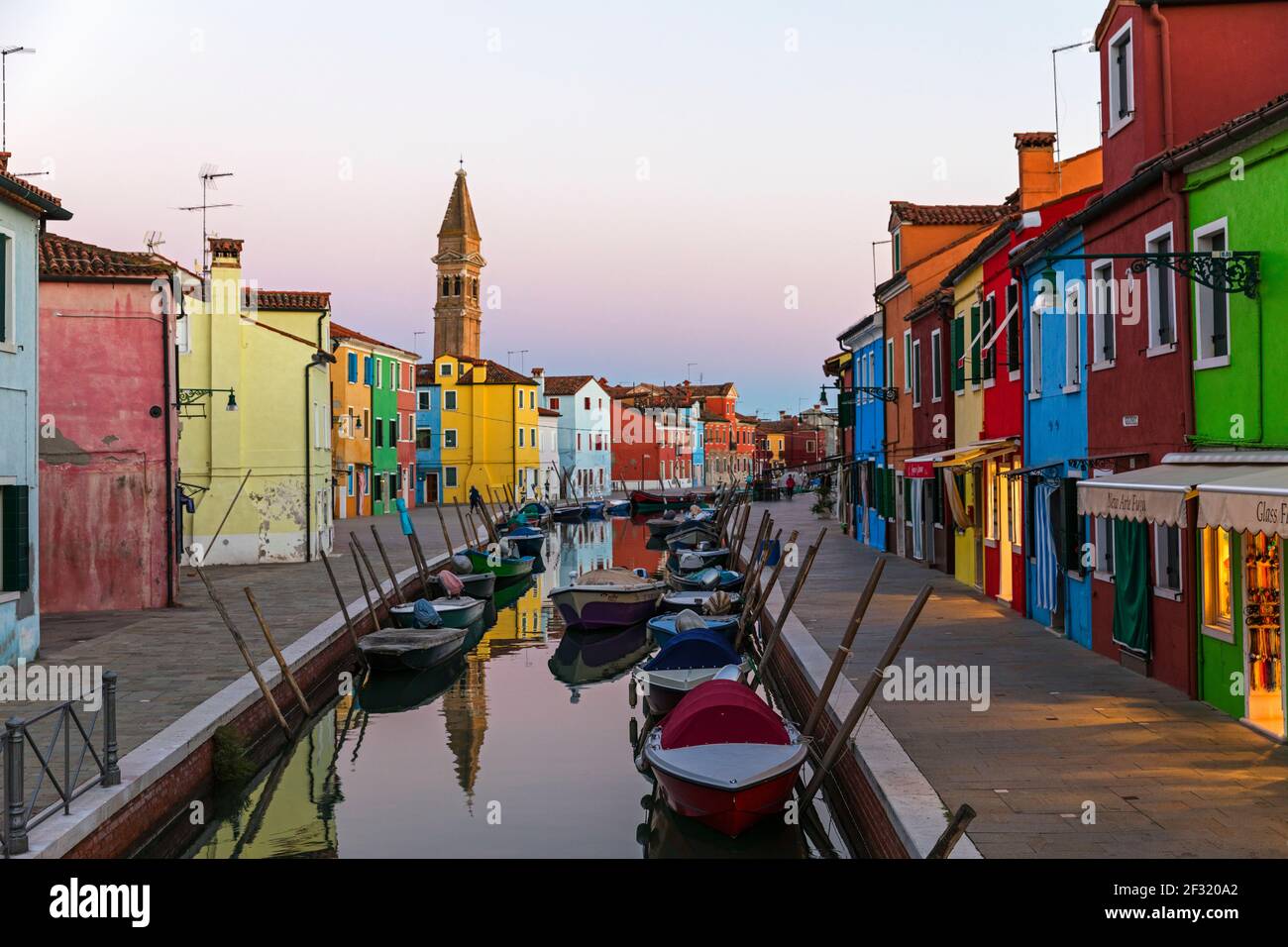 L'isola veneziana di Burano, barche ormeggiate su un canale, casa colorata e negozi al tramonto Foto Stock