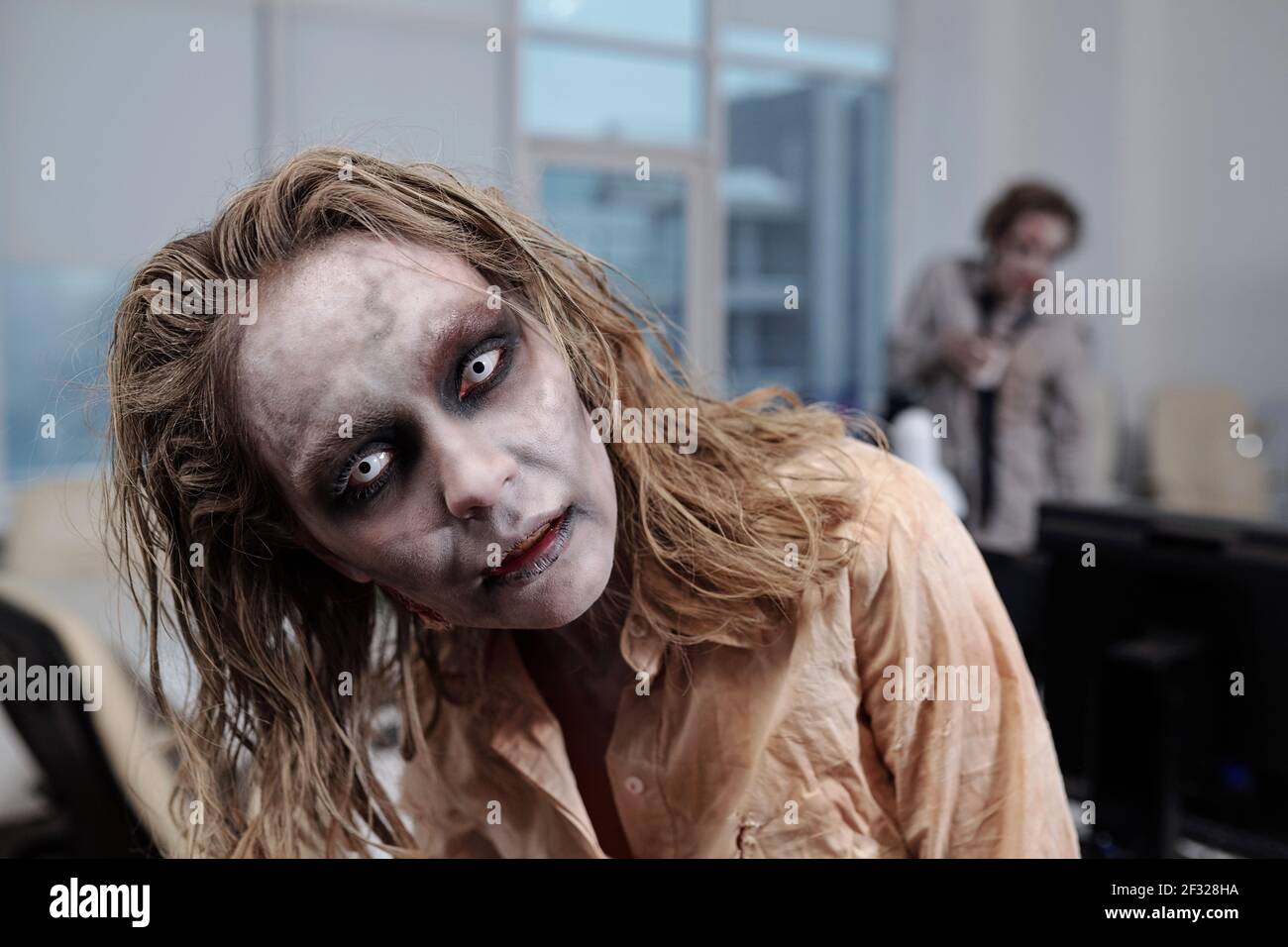Donna d'affari Zombie in piedi davanti alla macchina fotografica sullo sfondo di luoghi di lavoro e collega maschile morto in ufficio open space Foto Stock