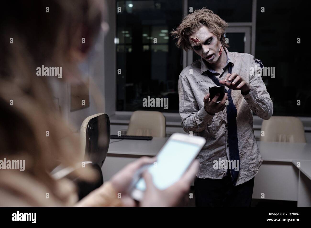 Uomo d'affari morto e spooky con greasepaint zombie su faccia e. scorrimento delle mani nello smartphone in ufficio a mezzanotte con femmina davanti Foto Stock