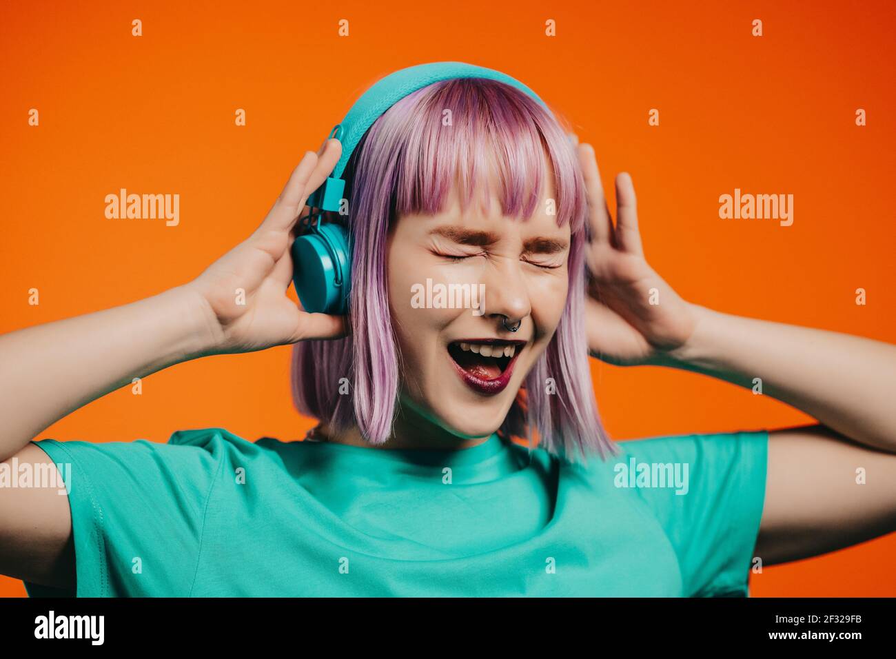 Donna eccezionale con capelli viola tinti che ascolta musica in cuffia e canta su sfondo arancione. Unica ragazza hipster danzare Foto Stock