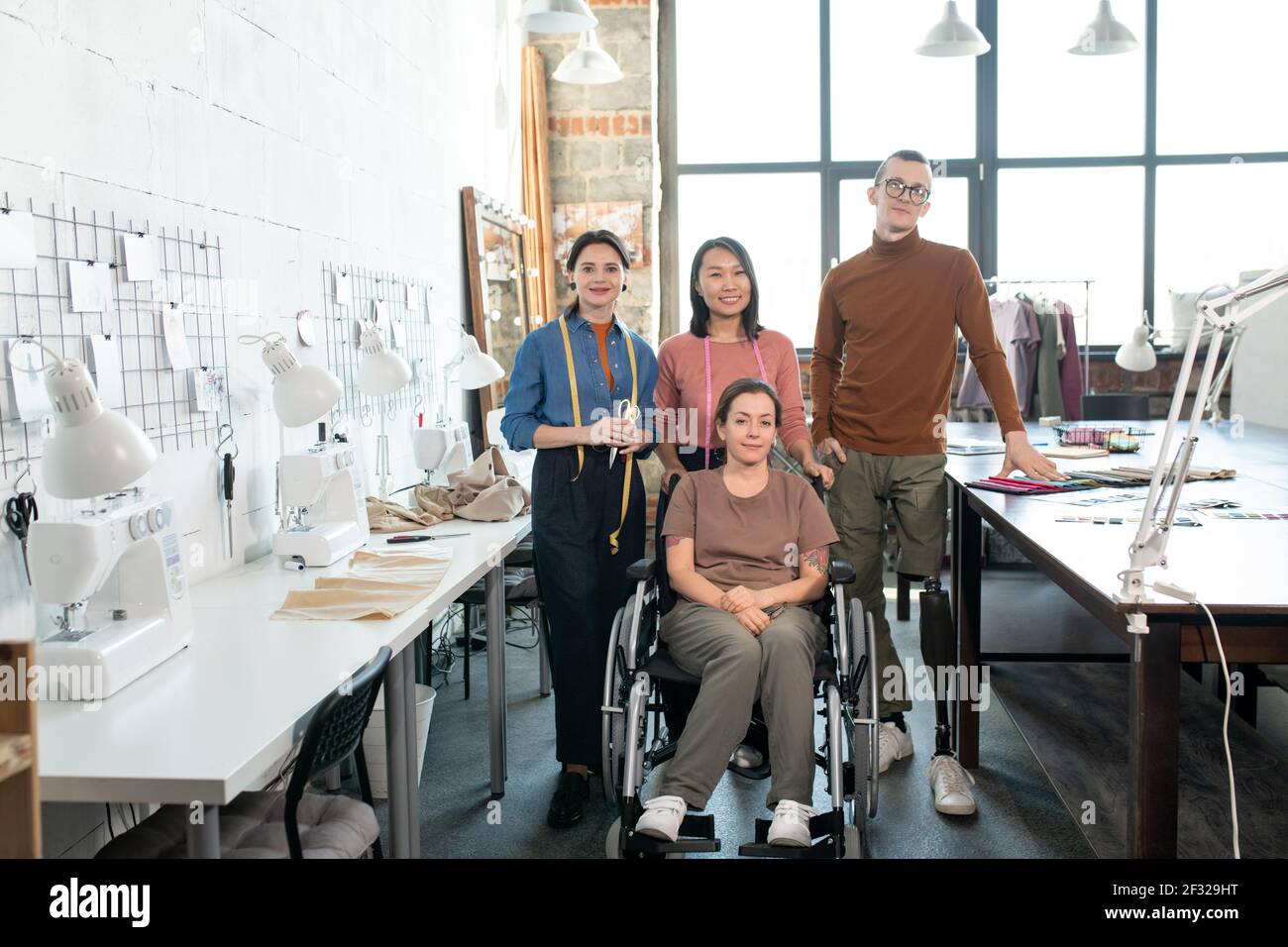 Team di stilisti contemporanei di successo e sarti in casualwear stare in piedi tra i tavoli con attrezzatura di lavoro Foto Stock
