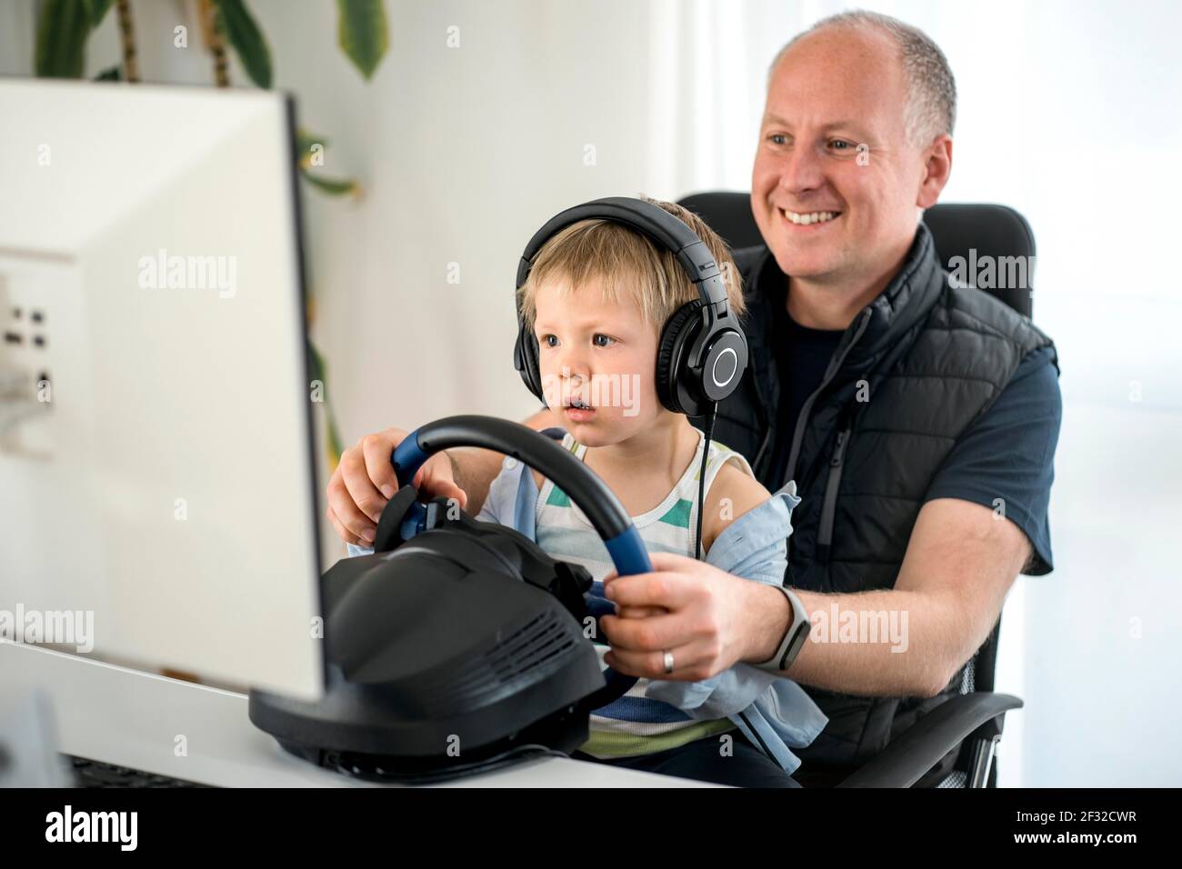 Ragazzo di tre anni che guarda con attenzione suo padre che gioca a correre gioco di automobile sul calcolatore usando un volante Foto Stock